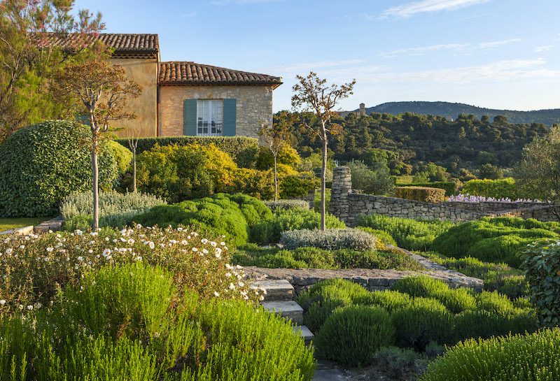 a provencal house to the left and gardens with herbs and shrubbery out front