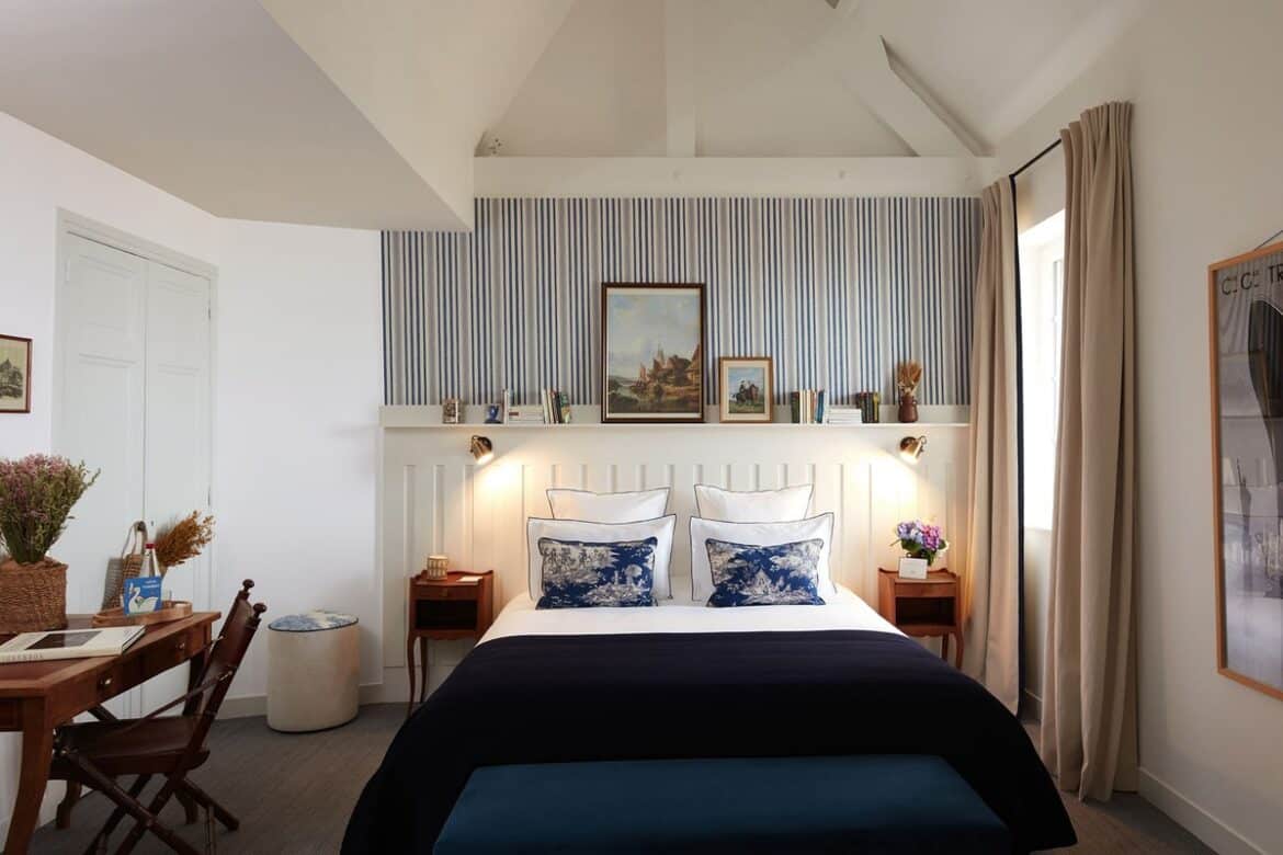Hotel Flaubert room interior with navy blue and white bedding, blue striped wallpaper, and vintage wood furniture.