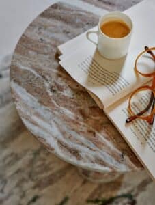 Calming cup of coffee, a book, and reading glasses on a marble table.