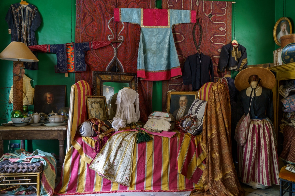 Eclectic wall of paintings, woven tapestries, and vintage clothes on display in a French antique store.