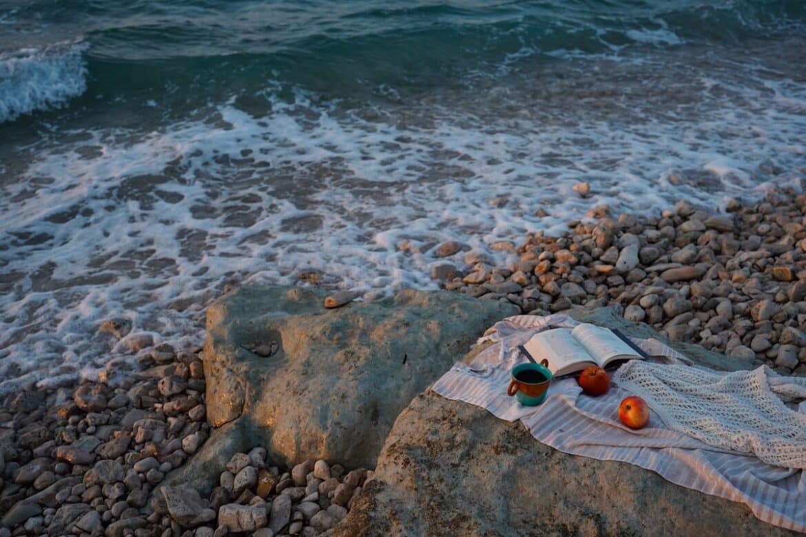 Open book, coffee, and fruit gathered on a blanket across a rock facing the incoming ocean tide.