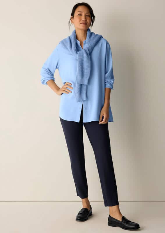 Blue Eileen Fisher cashmere sweater tied loosely over the shoulders with a blue button down, straight black pants, and loafers.