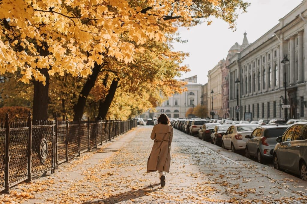 Woman in a long trench coat walking beneath trees in autumn.